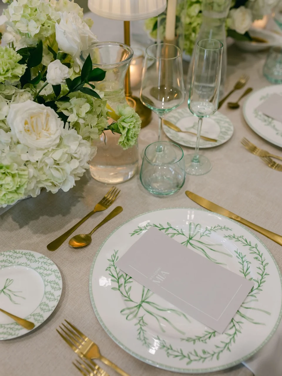Botanical Bow plates with green ribbon motifs, pale glassware and gold cutlery beside white flowers